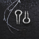 A close-up of two small metal tools with forked ends placed on a dark, textured background. To the left of the tools, thin white branches rest on the surface, adding contrast to the scene. The setting appears moody and detailed, reminiscent of displays for Kaya Movement's CALYPSO Minimalist Keyhole Ear Weights in 925 Silver, 2 gauge.