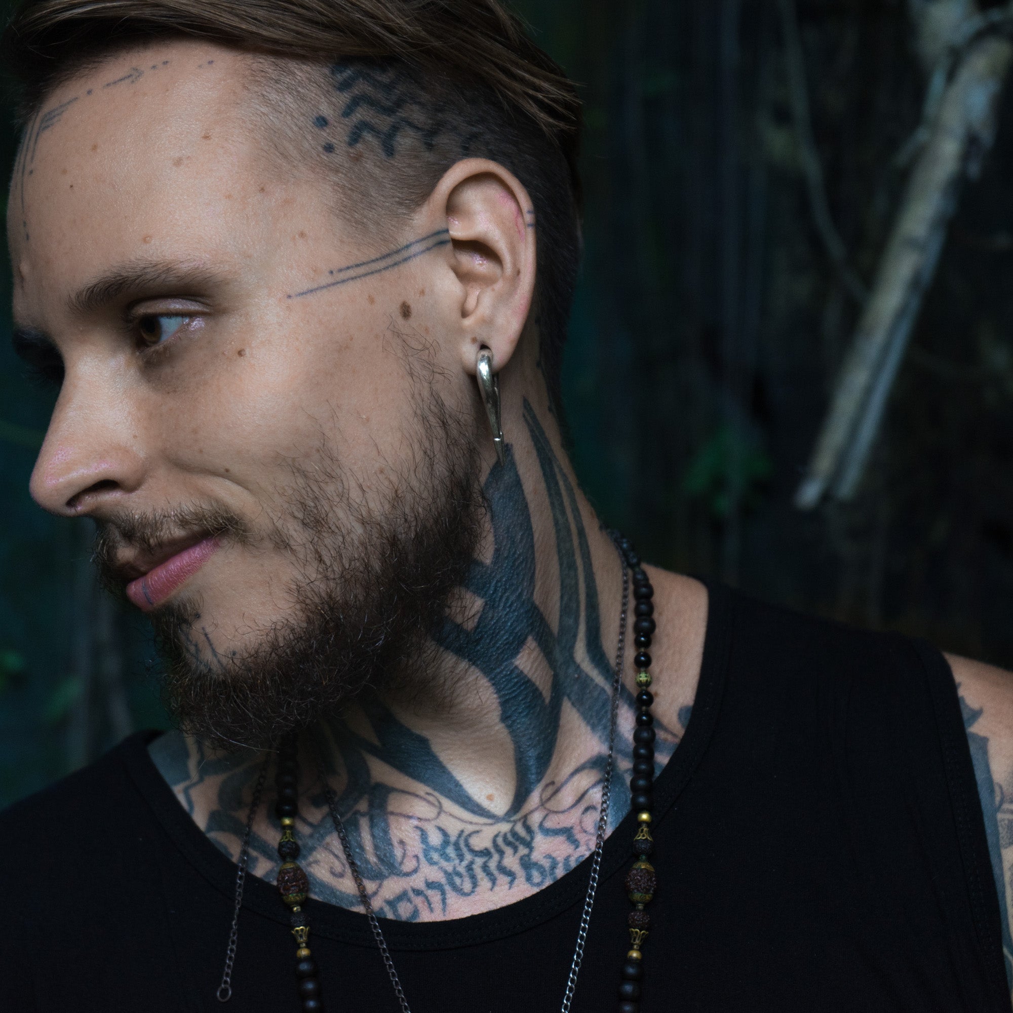 A person with a beard, ear piercing adorned with CALYPSO Minimalist Keyhole Ear Weights in 925 Silver by Kaya Movement, and extensive tattoos on the neck and face looks to the side. The individual is wearing a black tank top and a beaded necklace. The background appears dark and blurred, possibly depicting an outdoor setting.