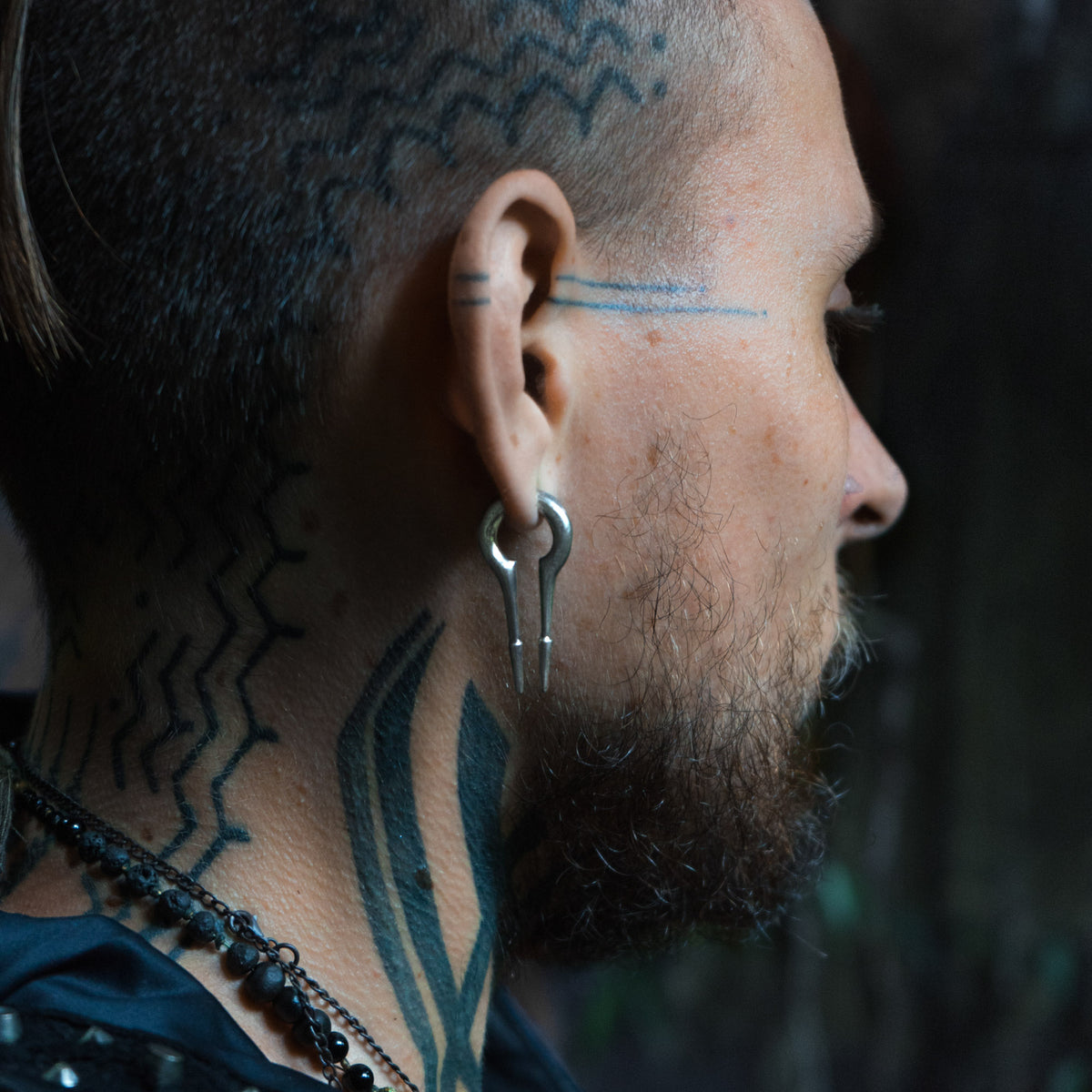 A close-up side profile captures a person with a shaved head adorned with intricate geometric tattoos on their scalp, neck, and face. They are wearing large, distinctive CALYPSO Minimalist Keyhole Ear Weights in 925 Silver by Kaya Movement and multiple necklaces. The background is blurred and dark.