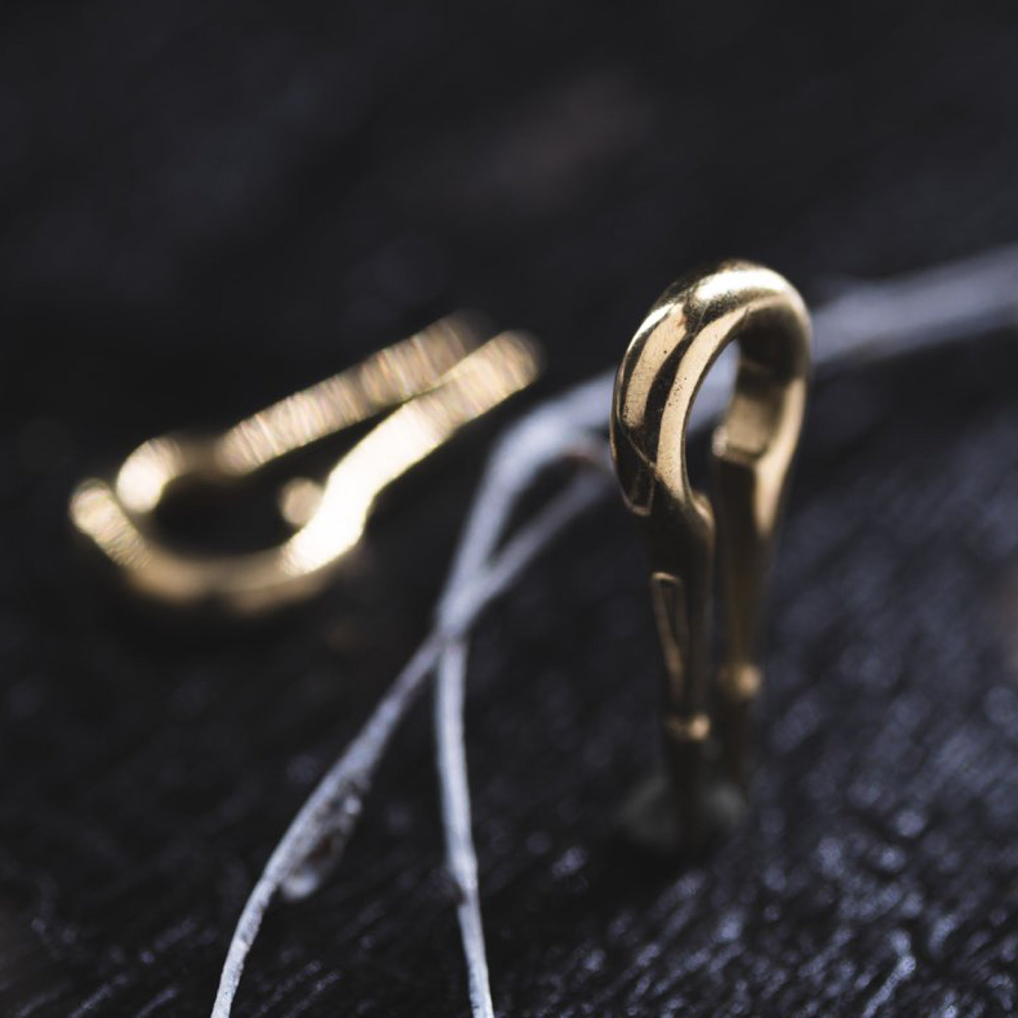Close-up image of the CALYPSO Minimalist Keyhole Ear Weights in Gold by Kaya Movement on a dark surface with thin strands of white thread or wire. Reminiscent of minimalist brass ear weights, the hooks are small, with one in focus at the front and another slightly blurred in the background.