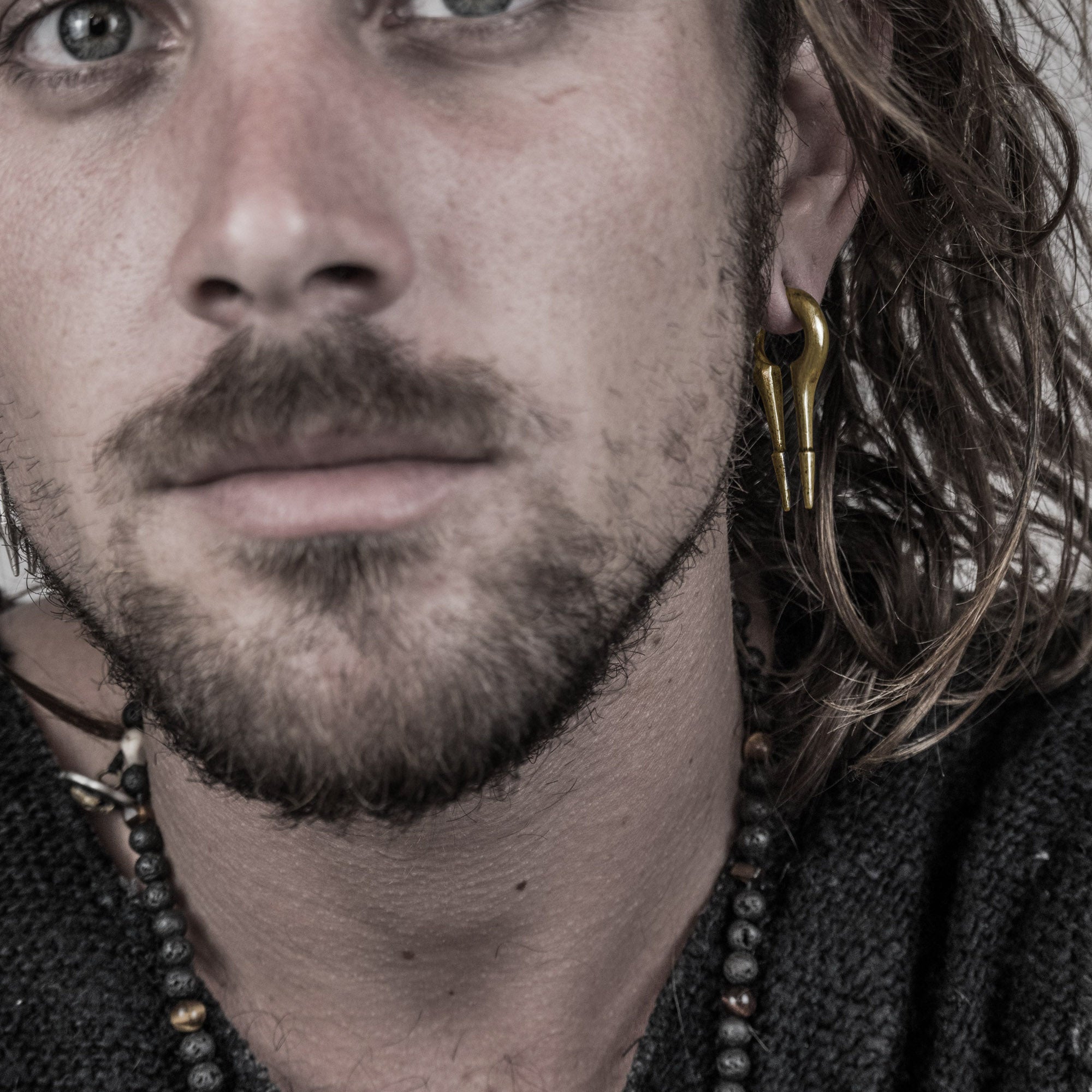 A close-up of a person with long wavy hair, a beard, and blue eyes. They are wearing a knit sweater, a beaded necklace, and Kaya Movement's CALYPSO Minimalist Keyhole Ear Weights in Gold (2 gauge). The background is out of focus, emphasizing their face.