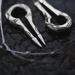 Close-up of the exquisitely crafted EXRO Biomechanical Cyborg Keyhole Ear Weights in 925 Silver by Kaya Movement, resting on a dark, textured surface. These 2 gauge ear weights feature a distinctive curved shape adorned with tribal-like designs etched into the silver, evoking the look of post-apocalyptic body jewelry.