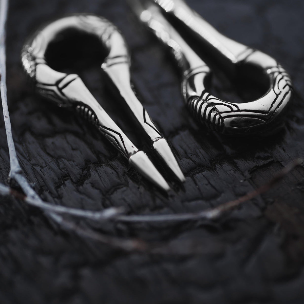Close-up of two intricately designed EXRO Biomechanical Cyborg Keyhole Ear Weights in Silver by Kaya Movement, resembling silver plier-like tools with a looped handle, set against a dark, textured surface. The ear weights feature ornate, linear patterns on their metallic surface, evoking the elegance of antique post-apocalyptic body jewelry.