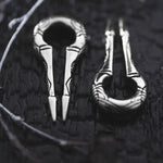 A close-up image of two intricately designed EXRO Biomechanical Cyborg Keyhole Ear Weights in 925 Silver by Kaya Movement placed on a dark, textured surface. The ear weights, reminiscent of post-apocalyptic body jewelry, have a circular loop on one end and tapered, pointed tines on the other, adorned with detailed engravings. The lighting emphasizes their metallic sheen.
