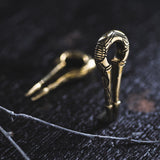 Two intricately designed EXRO Biomechanical Cyborg Keyhole Ear Weights in Gold Brass by Kaya Movement lie on a dark wooden surface. One ear weight is in focus, standing upright, while the other appears blurred in the background. Resembling unisex plug earrings, a twig rests in the foreground. The scene is softly lit, evoking an air of timeless elegance.