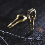 Two intricately designed EXRO Biomechanical Cyborg Keyhole Ear Weights in Gold Brass by Kaya Movement lie on a dark wooden surface. One ear weight is in focus, standing upright, while the other appears blurred in the background. Resembling unisex plug earrings, a twig rests in the foreground. The scene is softly lit, evoking an air of timeless elegance.