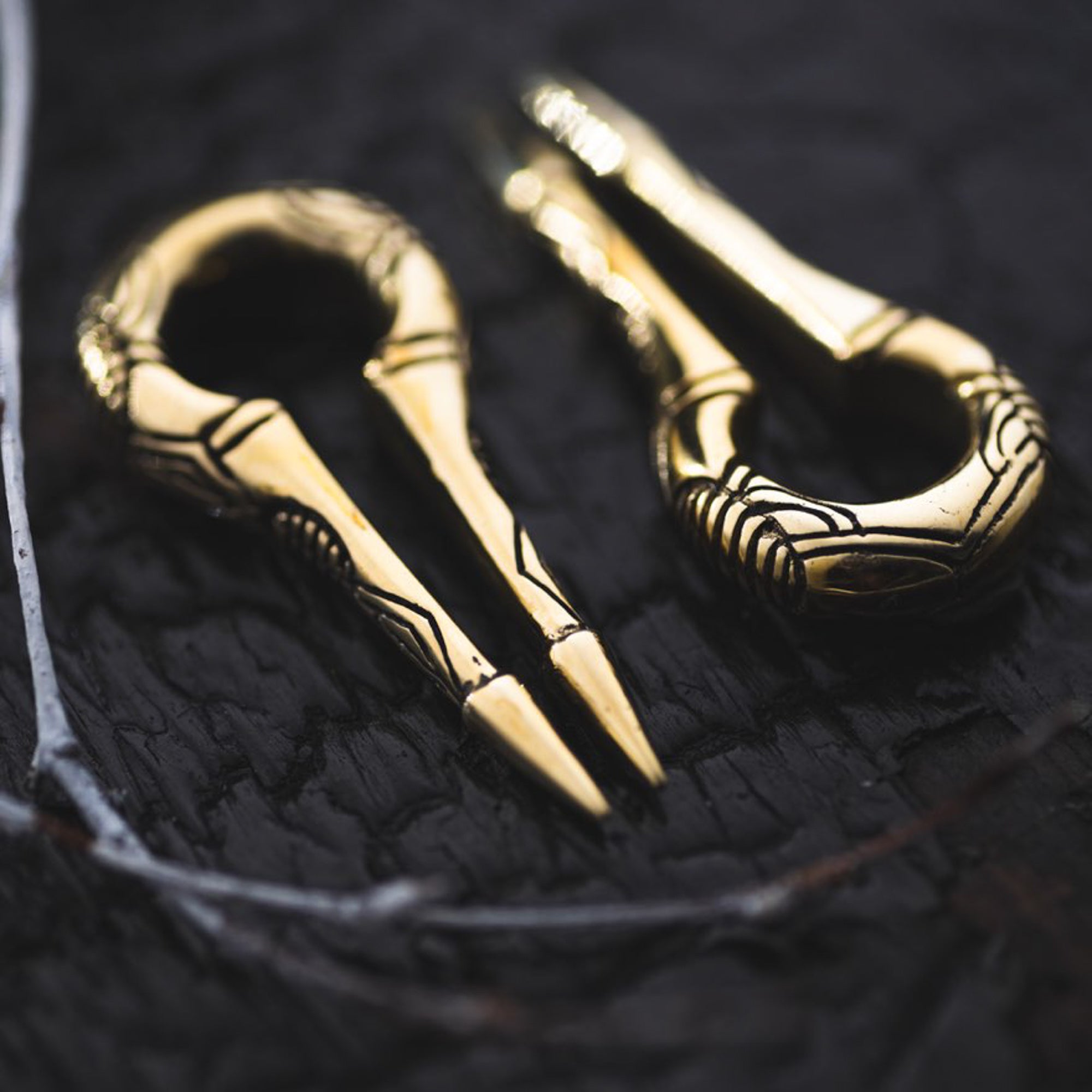 Close-up of two EXRO Biomechanical Cyborg Keyhole Ear Weights in Gold Brass by Kaya Movement on a dark surface. The ear weights showcase a simple, elegant design with linear engravings, reminiscent of unisex plug earrings. The surface texture is rough and slightly reflective, contrasting with the smooth, shiny gold brass ear weights.