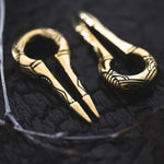 Close-up of two EXRO Biomechanical Cyborg Keyhole Ear Weights in Gold Brass by Kaya Movement on a dark surface. The ear weights showcase a simple, elegant design with linear engravings, reminiscent of unisex plug earrings. The surface texture is rough and slightly reflective, contrasting with the smooth, shiny gold brass ear weights.