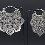A pair of intricately designed ASA Oversized Asa No Ha Mandala Disc Earrings in Silver from Kaya Movement hangs on black display stands against a dark background. The earrings feature a geometric, lace-like mandala pattern with a combination of hexagonal and floral motifs, accentuated by an Asanoha motif that highlights their detailed craftsmanship.