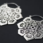 Close-up image of two ornately designed ASA Oversized Asa No Ha Mandala Disc Earrings in Silver by Kaya Movement, featuring intricate geometric patterns laid on a dark background. The 16 gauge earrings showcase an Asanoha motif with multiple layers and shapes, creating an elaborate, delicate appearance.
