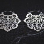Close-up of a pair of Kaya Movement's ASA Oversized Asa No Ha Mandala Disc Earrings in Silver (16 gauge) with intricate, mandala-shaped tribal designs featuring delicate cut-out patterns, set against a dark, textured background. The design blends geometric and floral elements to create a symmetrical, ornate appearance.