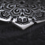 Close-up image showcasing the metallic, intricately designed geometric pattern on a dark surface. The design features symmetrical shapes and sharp edges arranged in a mandala-like structure, reminiscent of an Asanoha motif. The surface is dark and textured, providing contrast to the reflective silver ASA Oversized Asa No Ha Mandala Disc Earrings by Kaya Movement, crafted in 16 gauge silver.