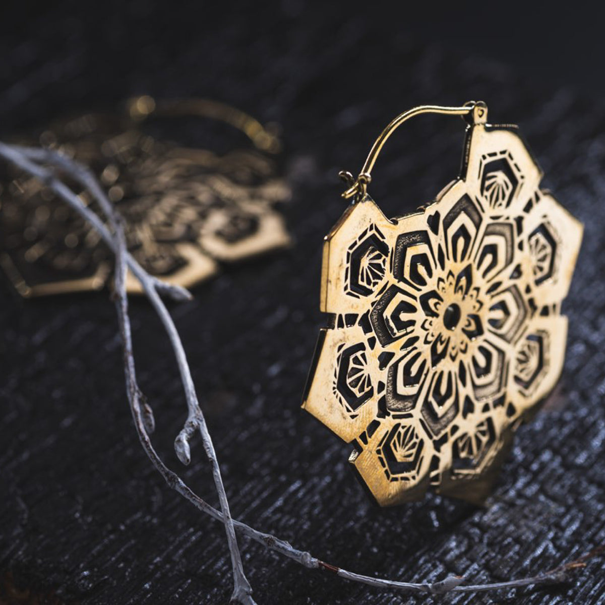 Close-up of an intricate ASA Oversized Asa No Ha Mandala Disc Earring in Gold by Kaya Movement, lying on a textured dark surface. Thin, dried branches are partly visible in the background, adding a rustic touch to the composition.