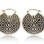 The AREVALO Shipibo Oversized Disc Hoop Earrings in Gold by Kaya Movement feature circular discs adorned with intricate black geometric designs on a white background. The pattern, including diamond shapes and crosses reminiscent of Sacred geometry Shipibo patterns, is attached to 14 gauge gold hoops.