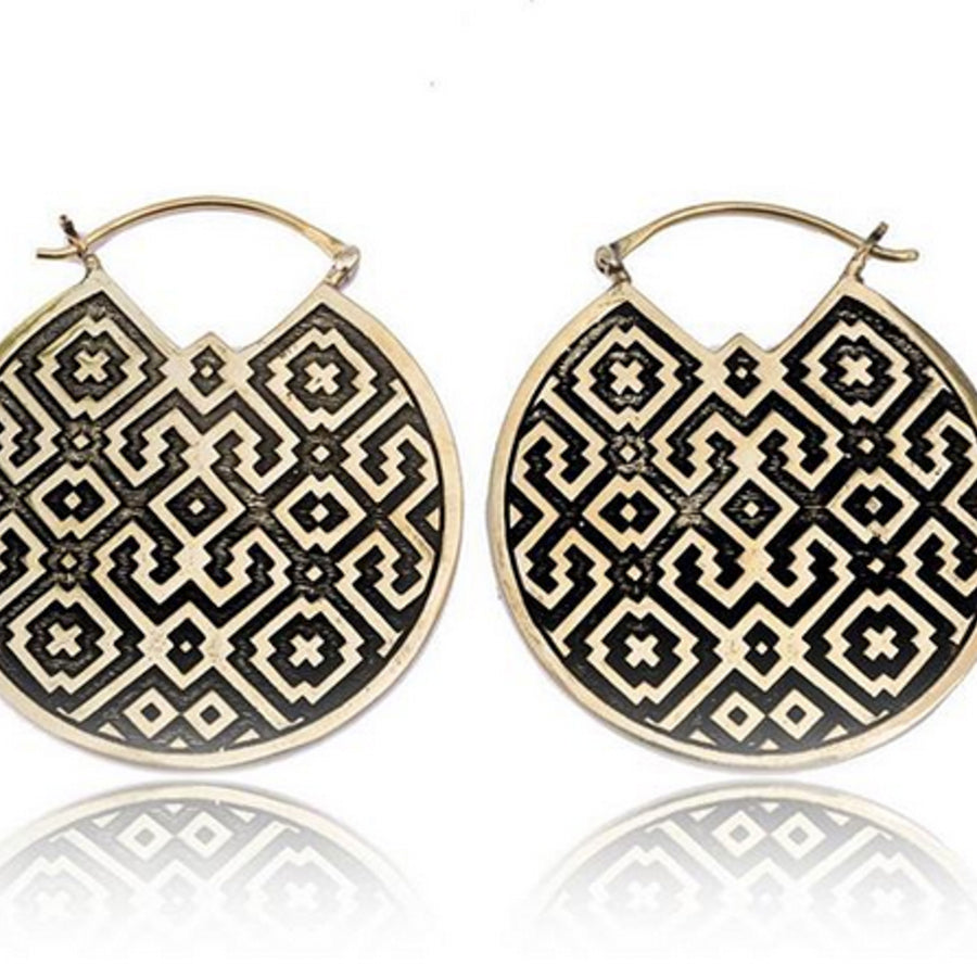 A pair of circular golden earrings with a distinctive black Shipibo-conibo pattern, presented on a reflective surface that creates a symmetrical image of their intricate design. The earrings have an elegant latch and are positioned side by side, their artistic detail symbolizing a deep cultural heritage.