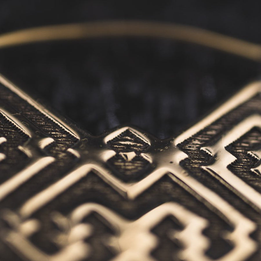 A macro photograph capturing the detailed texture and pattern of a piece of jewelry, showcasing the sharp contrast between the gleaming golden edges and the dark, intricately embossed designs on its surface. The depth of field is shallow, bringing the central part of the pattern into focus against the softly blurred surroundings.