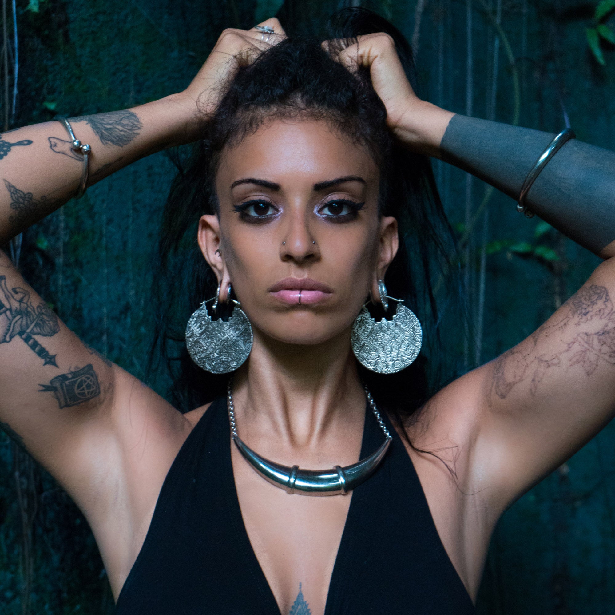 A woman with PANCHA Oversized Tribal Shipibo Disc Earrings in Silver by Kaya Movement and a silver necklace stands with her hands behind her head. She has long black hair, tattoos on her arms, and wears a black halter top. The background is dark with a greenish hue, possibly a mossy or natural setting.