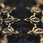 Close-up of an intricately designed metallic object with a symmetrical pattern, reminiscent of Mozika Oversized Gothic Disc Earrings in Gold Brass by Kaya Movement. The aged metal has a dark, reflective surface in the background. The pattern features interconnected shapes, possibly floral or geometric, with a golden hue.