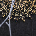 A close-up image of intricately designed gold mandala against a dark background evokes the elegance of Kaya Movement’s Mozika Oversized Gothic Disc Earrings in Gold Brass | 16 gauge. The delicate patterns feature loops and teardrop shapes, with thin white branches intersecting the mandala, adding contrast to the detailed artwork.