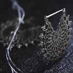 A close-up shot of an intricately designed MOZAIKA Oversized Gothic Disc Earring in Silver by Kaya Movement resting on a dark, slightly textured surface. The earring features a complex web-like pattern, and a thin branch with similarly detailed patterns can be seen in the background.