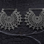 Displayed on a dark, textured background are the MOZAIKA Oversized Gothic Disc Earrings in Silver by Kaya Movement. These earrings feature an intricate, lace-like pattern with delicate interwoven metalwork and small hoops at the top for wearing.