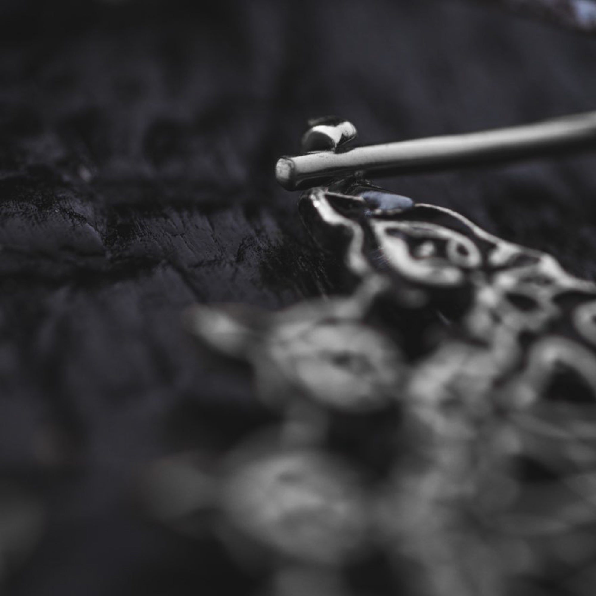 Close-up of the MOZAIKA Oversized Gothic Disc Earrings in Silver by Kaya Movement, showcasing the intricate and ornate designs. The earrings, made with 16 gauge metal, are lying on a dark, textured surface. The image artistically focuses on a fine detail at the top part of the silver piece, leaving the rest slightly blurred.