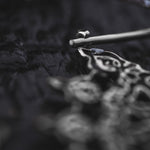 Close-up of the MOZAIKA Oversized Gothic Disc Earrings in Silver by Kaya Movement, showcasing the intricate and ornate designs. The earrings, made with 16 gauge metal, are lying on a dark, textured surface. The image artistically focuses on a fine detail at the top part of the silver piece, leaving the rest slightly blurred.
