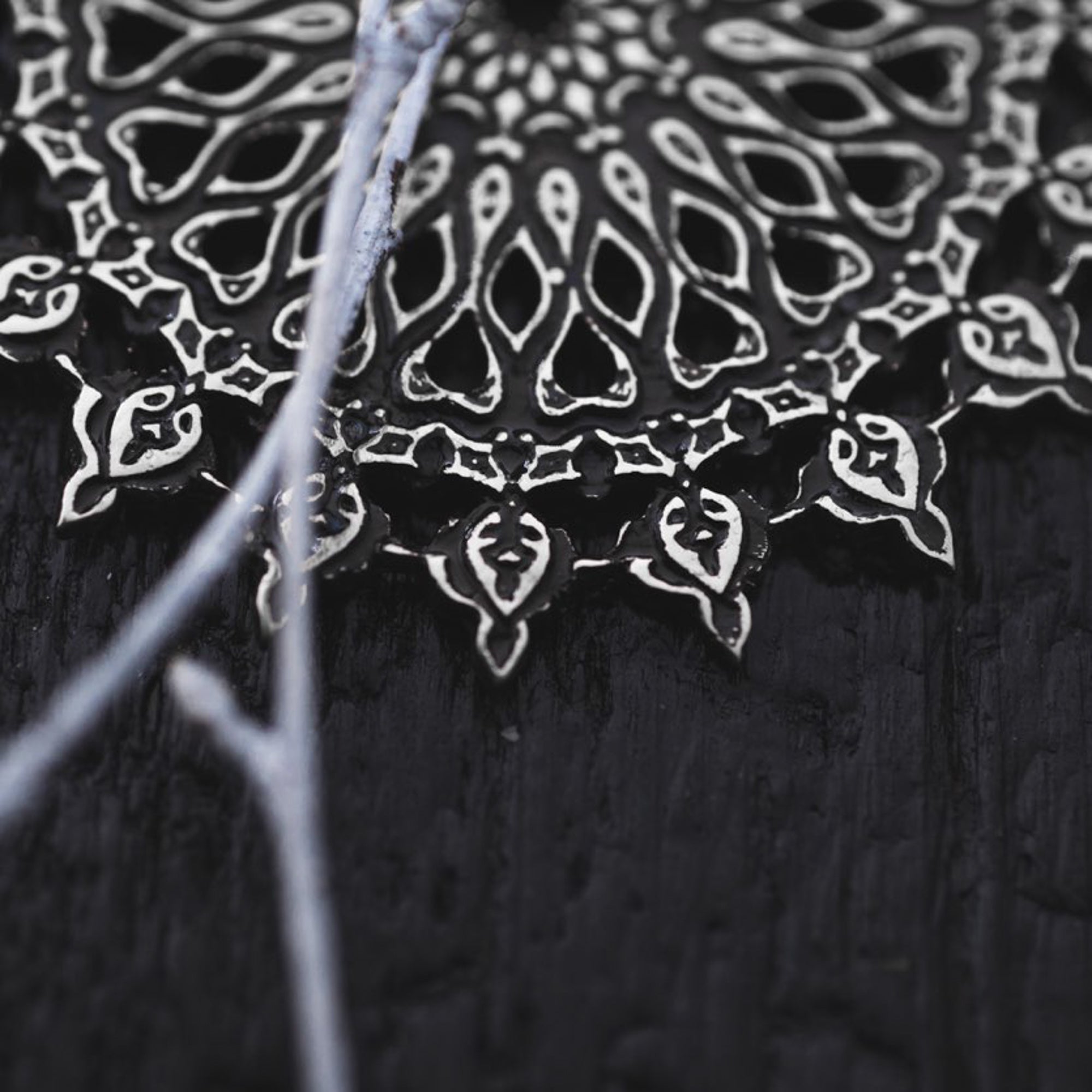 A close-up view reveals an intricately designed black and white mandala adorned with detailed, symmetrical motifs that resemble petals or leaves, all radiating from a central point. Interspersed light-colored threads add contrast, reminiscent of the delicate lines on Kaya Movement's MOZAIKA Oversized Gothic Disc Earrings in Silver (16 gauge).
