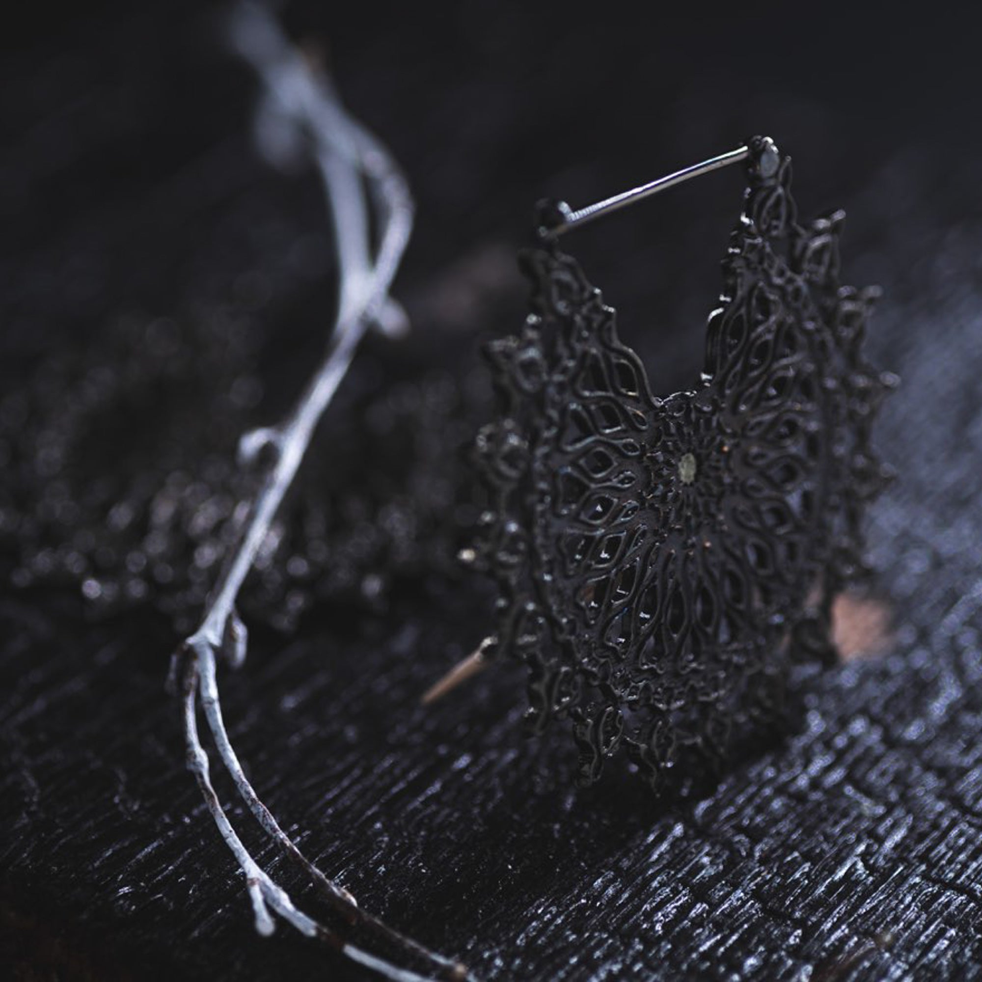 A delicate, intricately designed pair of MOZAIKA Oversized Gothic Disc Earrings in Black by Kaya Movement rests on a dark wooden surface. The intricate pattern is complex and lacy, embodying the elegance of these 16-gauge Gothic disc earrings. Nearby, a thin white twig with a few branches adds contrasting texture to the moody, dimly-lit scene.