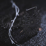 A delicate, intricately designed pair of MOZAIKA Oversized Gothic Disc Earrings in Black by Kaya Movement rests on a dark wooden surface. The intricate pattern is complex and lacy, embodying the elegance of these 16-gauge Gothic disc earrings. Nearby, a thin white twig with a few branches adds contrasting texture to the moody, dimly-lit scene.