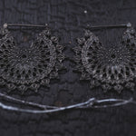 Close-up of a pair of MOZAIKA Oversized Gothic Disc Earrings in Black by Kaya Movement, intricately designed with a circular, lace-like pattern. They rest on a textured black surface, with a small portion of a silver chain visible below them. The intricate design gives an elegant and detailed appearance to these 16 gauge dark metallic round hoops.