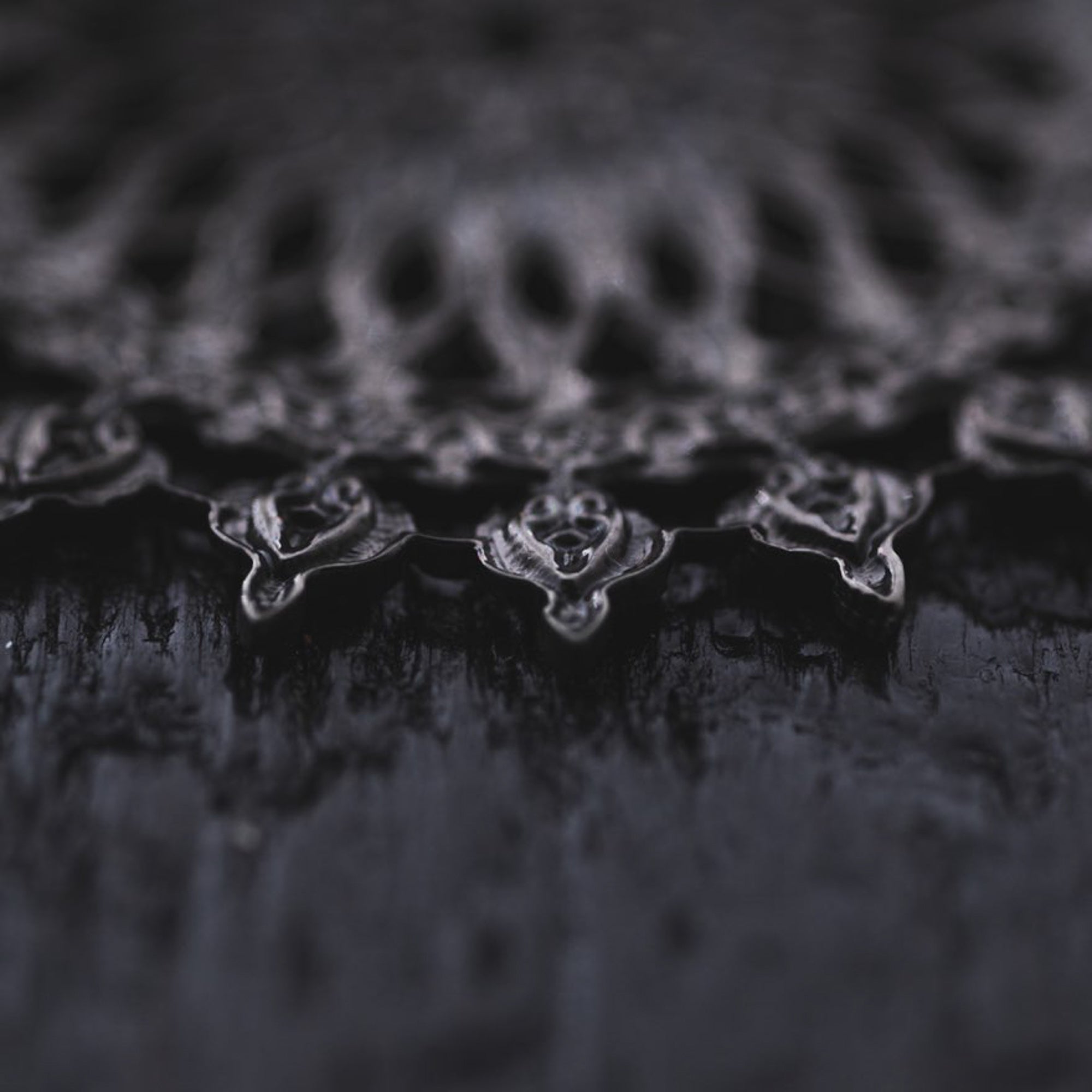A close-up image presents the intricate design of the MOZAIKA Oversized Gothic Disc Earrings in Black by Kaya Movement. The delicate lace-like pattern against a subtly blurred black textured background highlights the 16 gauge earrings' fine details and symmetrical shapes, evoking a sense of depth and complexity. The overall tone is moody and artistic, capturing the essence of these striking accessories.