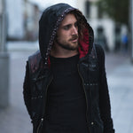 A man with shoulder-length hair and a beard stands outdoors on a street, wearing the VENOM Men's Leather Hooded Vest Jacket with red lining from Kaya Movement, resembling a punk rock biker jacket. He looks to the left with a thoughtful expression. The background is slightly blurred, showing an urban setting with buildings and a person in the distance.