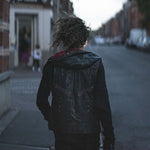 A person with curly hair, wearing the VENOM Men's Leather Hooded Vest Jacket by Kaya Movement, adorned with studded details, walks down an empty street in the evening. The background is blurred, featuring buildings and a few people near a shop entrance on the left.