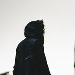 A silhouette of a person wearing the Kaya Movement VENOM Men's Leather Hooded Vest Jacket with its distinctive red lining stands against a bright, overcast sky. The hood covers their head, and their face is not visible, creating an air of mystery and contemplation.