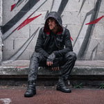 A person in a VENOM Men's Leather Hooded Vest Jacket with Red Lining by Kaya Movement and fingerless gloves sits on a ledge against a graffiti-covered wall. They wear dark pants, rugged boots, and sport a contemplative expression. The gritty, urban surroundings enhance the moody, street-style aesthetic.