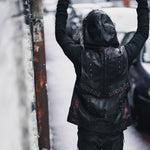 A person wearing the VENOM Men's Leather Hooded Vest Jacket with red lining by Kaya Movement stands with their back to the camera, raising their arms up in an urban environment next to a weathered wall and parked cars on a wet street.