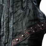 Close-up of the VENOM Men's Leather Hooded Vest Jacket by Kaya Movement, showcasing black leather with textured stitching and a decorative strip. The strip is adorned with round, metallic studs and features a red, perforated layer underneath. The leather has a wrinkled and worn texture, adding to its rugged, punk rock look.
