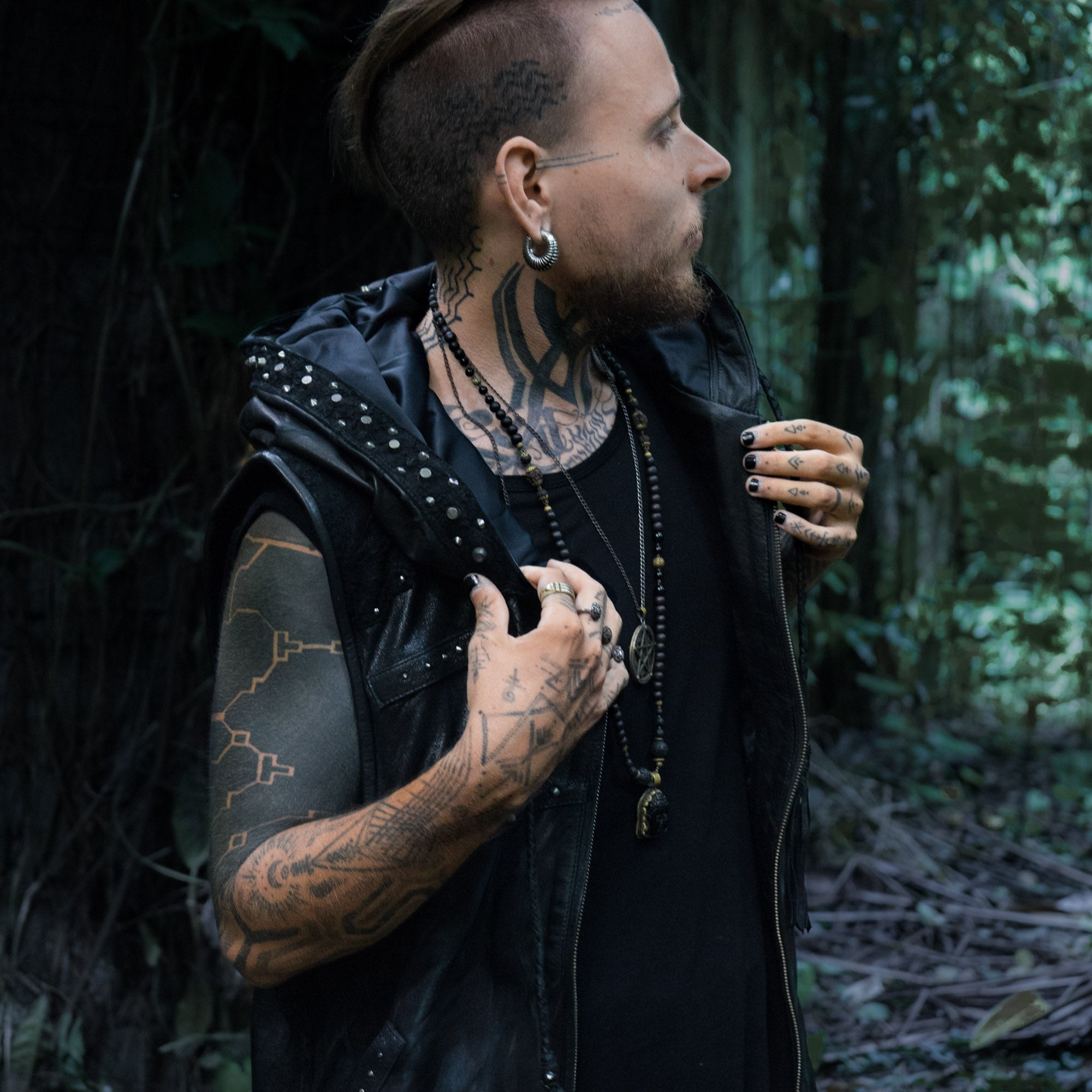 A person with tattoos on their head, neck, and arms stands outdoors in front of a forest backdrop, looking to the side. They are wearing the VENOM Men's Leather Hooded Vest Jacket by Kaya Movement, a sleeveless black leather hoodie adorned with studs. They have gauges in their ears and are holding the collar of this custom leather good.