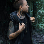 A person with tattoos on their head, neck, and arms stands outdoors in front of a forest backdrop, looking to the side. They are wearing the VENOM Men's Leather Hooded Vest Jacket by Kaya Movement, a sleeveless black leather hoodie adorned with studs. They have gauges in their ears and are holding the collar of this custom leather good.