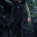 A person wearing the VENOM Men's Leather Hooded Vest Jacket by Kaya Movement, a black shirt, and ripped black jeans stands outside against a backdrop of dense, dark green foliage. They have multiple tattoos on their arms and are adorned with necklaces. Their face is not visible.