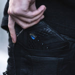 A person is placing or retrieving a Kaya Movement's AMONI Black Stud Bi-Fold Flip Leather Wallet with a Labradorite emblem from a pocket. The focus is on the wallet and their fingers, with the person's clothing and background blurred.
