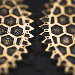 Close-up image of a dark surface with intricate, honeycomb-like gold patterns resembling the detailing on the Kaya Movement ILLUSION Oversized Cyborg Sci Fi Disc Earrings in Gold. The geometric shapes have a slightly textured appearance, forming two symmetrical designs that do not touch but align closely. The details are sharp, highlighting the 14-gauge metallic finish.