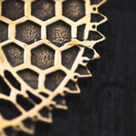 Close-up view of the ILLUSION Oversized Cyborg Sci Fi Disc Earrings in Gold by Kaya Movement against a dark background. The earrings, resembling post-apocalyptic jewelry, appear to be made of a golden material with irregular edges, highlighting their hexagonal pattern. The background texture is rough and black.
