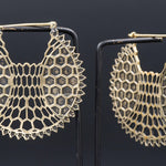 A pair of ILLUSION Oversized Cyborg Sci Fi Disc Earrings in Gold by Kaya Movement hangs against a dark background. The gold earrings showcase a honeycomb pattern forming a crescent shape, with tiny geometric cutouts creating a delicate, lace-like appearance. They are supported by thin, black stands, echoing the elegance of post-apocalyptic jewelry.