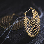 Close-up view of the ILLUSION Oversized Cyborg Sci Fi Disc Earrings in Gold by Kaya Movement against a dark, blurred background. The earring features a hexagonal, honeycomb-like pattern and a crescent shape, with a delicate wire looping through it adding detail to the composition, exuding an almost post-apocalyptic jewelry vibe.