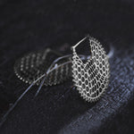 A close-up of a pair of intricate, oversized disc-shaped earrings named "ILLUSION" by Kaya Movement. These 14 gauge silver earrings feature a delicate, hexagonal pattern that gives them a textured, lace-like appearance. Embodying subtle sci-fi vibes, they are placed on a dark wooden surface, creating a striking contrast with their metallic sheen.
