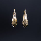 A close-up of the PROMETHEUS Medium Biomechanical Hoop Earrings in Gold by Kaya Movement against a dark background. The brass-colored earrings feature a detailed and ornate pattern, resembling a vintage or tribal style. The dark background emphasizes the metallic sheen of these 18-gauge sci-fi statement earrings.