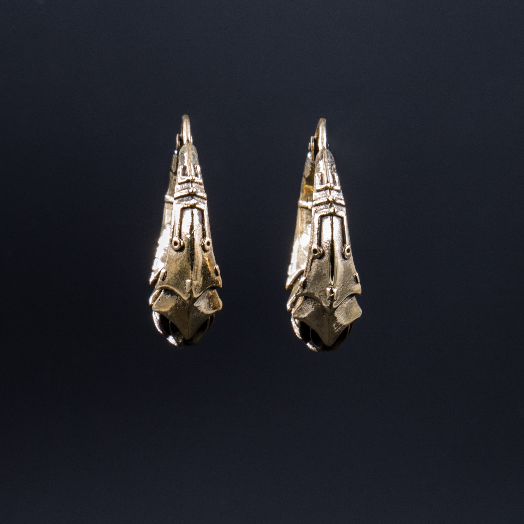 A close-up of the PROMETHEUS Medium Biomechanical Hoop Earrings in Gold by Kaya Movement against a dark background. The brass-colored earrings feature a detailed and ornate pattern, resembling a vintage or tribal style. The dark background emphasizes the metallic sheen of these 18-gauge sci-fi statement earrings.