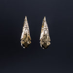 A close-up of the PROMETHEUS Medium Biomechanical Hoop Earrings in Gold by Kaya Movement against a dark background. The brass-colored earrings feature a detailed and ornate pattern, resembling a vintage or tribal style. The dark background emphasizes the metallic sheen of these 18-gauge sci-fi statement earrings.